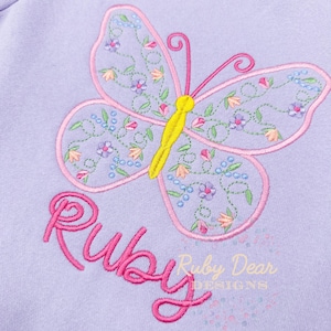 May include: A lavender sweatshirt with a pink butterfly embroidered on the front. The butterfly has yellow accents and is surrounded by small pink and blue flowers. The word "Ruby" is embroidered in pink below the butterfly.