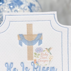 May include: White fabric with a light blue stitched border and a cross design. The cross is tan with light blue draping. The words "He Is Risen" are embroidered in light blue. A floral and hummingbird logo is in the upper right corner.