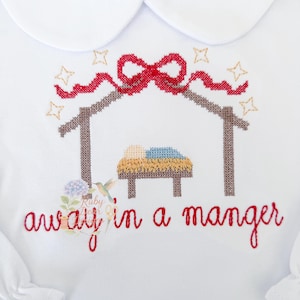 May include: White long-sleeved shirt with a Peter Pan collar. The shirt features a cross-stitch design of a manger scene with a red bow and the words "away in a manger" in red script. Gold stars are embroidered around the manger.