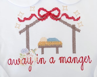 Könnte beinhalten: Weißes Langarmhemd mit einem Bubikragen. Das Hemd zeigt ein Kreuzstichdesign einer Krippenszene mit einer roten Schleife und den Worten "away in a manger" in roter Schrift. Goldene Sterne sind um die Krippe herum gestickt.