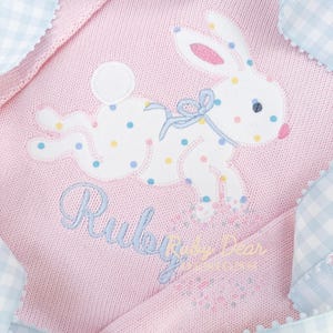 Puede incluir: Suéter de punto rosa para bebé con un conejito blanco bordado en la parte delantera. El conejito tiene un lazo azul y lunares de colores. El suéter tiene un ribete de cuadros azules y blancos y el texto "Ruby Baby Dear" está bordado en la parte inferior.