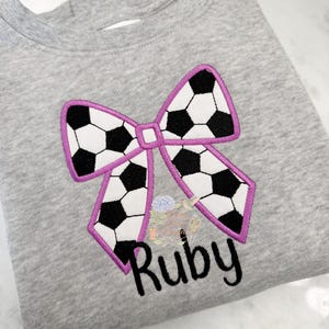 May include: A gray sweatshirt featuring a soccer ball bow design with a purple outline. The bow is made of black and white soccer ball patterns. The name "Ruby" is embroidered in black script below the bow.