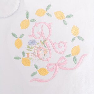 May include: White garment with a scalloped collar, embroidered with a pink letter "R" surrounded by yellow lemons and green leaves. A pink bow and the text "Ruby Dear Designs" are also embroidered on the garment.