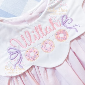 May include: A pink and white baby dress with a white embroidered collar featuring a donut garland and the name "Allie" in pink script. The collar has a purple bow on each side.
