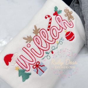 Könnte beinhalten: Weißer Weihnachtspullover mit rosa gesticktem Schriftzug "Willa" und verschiedenen weihnachtlichen Stickereien, darunter ein Zuckerstange, ein Lebkuchenmann, ein Weihnachtsbaum, ein Geschenk, ein Rentier und ein Weihnachtsbaumschmuck.