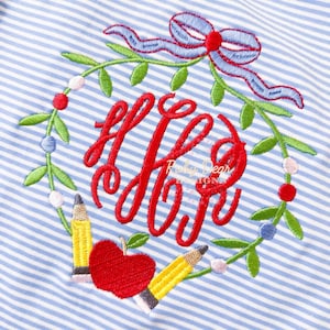 May include: A blue and white striped shirt with an embroidered wreath design. The wreath is decorated with red apples, yellow pencils, and green leaves. The text "H.P.S." is embroidered in red within the wreath.