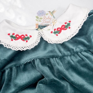 Può includere: Un vestito in velluto a coste verde acqua con colletto bianco smerlato. Il colletto è ricamato con un fiocco rosso e agrifoglio verde. L'abito è un capo festivo, perfetto per le festività natalizie.