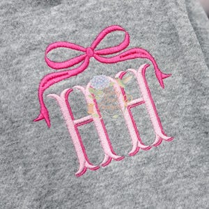 May include: Gray fabric with a pink embroidered bow and the letters "HH" in a decorative font. The embroidery is detailed, with a floral and hummingbird design in the center of the letters.
