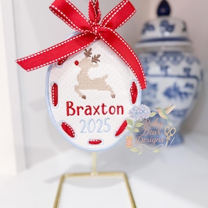 May include: An embroidered Christmas ornament in an oval shape, featuring a reindeer design and the name "Braxton" with the year 2025. The ornament has a light blue border, red accents, and is topped with a red ribbon. It is displayed on a gold stand.