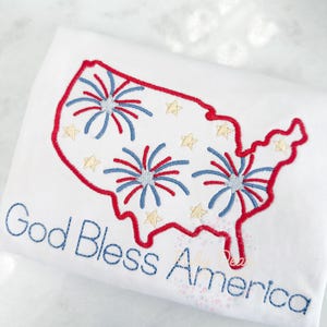 Könnte beinhalten: Weißes T-Shirt mit einer gestickten Umrandung der Vereinigten Staaten mit roten, weißen und blauen Feuerwerkskörpern im Inneren. Der Text "God Bless America" ist unterhalb der Umrandung gestickt.