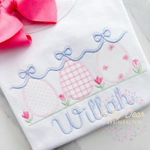 May include: A white onesie with a blue and pink embroidered design of three Easter eggs with bows and flowers. The onesie is personalized with the name "Willa" in blue embroidery.