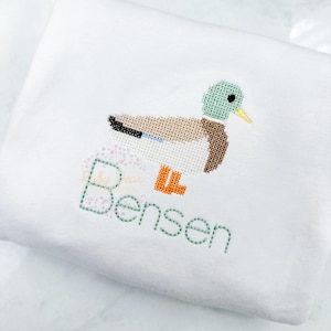 Puede incluir: Un body blanco para bebé con un pato bordado en verde, marrón y naranja, y el nombre "Bensen" bordado debajo.