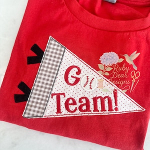 Könnte beinhalten: Rotes T-Shirt mit einem Wimpel-Design mit der Aufschrift "Go Team!" in roter Schrift. Der Wimpel hat eine Baseball-Grafik und ein Vichy- und Tupfenmuster. Das Shirt hat auch ein Blumen- und Kolibri-Design.