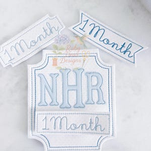 Op de afbeelding: Geborduurde baby-mijlpaalmarkeringen in wit met blauwe stiksels. De set bevat een grote plaquette met de initialen "NHR" en de tekst "1 Month", plus twee kleinere "1 Month" banners. De achtergrond heeft een subtiel stippenpatroon.