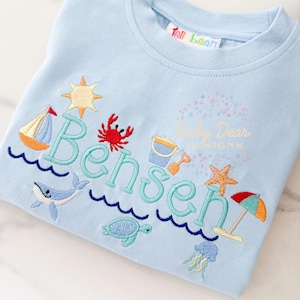 Może przedstawiać: Jasnoniebieska koszulka z haftem o tematyce plażowej. Zawiera żaglówkę, kraba, słońce, wieloryba, żółwia, wiaderko z piaskiem, rozgwiazdę, parasol i imię "Bensen" w kolorze turkusowym. Koszulka zawiera również napis "Baby Dear Designs".