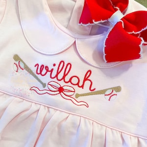 May include: Pink baby dress with a white collar and a red bow. The dress has an embroidered design of two baseball bats, a baseball, a red bow, and the name "willah".