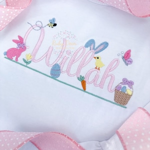 May include: A white babygrow with pink and blue embroidered Easter-themed designs. The babygrow is personalised with the name "Willa" in pink script. The design includes a bunny, chick, butterfly, and a basket of eggs.