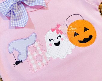 May include: A pink short-sleeved shirt with a purple and white gingham bow. The shirt has an applique design of a ghost, a spider, a pumpkin, and the word "Boo" in pink and white gingham.