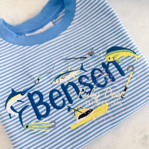 May include: A blue and white striped t-shirt with a blue collar. The shirt features embroidered fishing-themed designs, including fish, hooks, and a boat, along with the name "Bensen" and "Ruby Dear Designs".