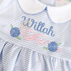 May include: Close-up of a baby's dress with a blue and white striped pattern. The dress features a white collar and is embroidered with the name "Willah" in blue script, along with a pink bow and blue floral accents.