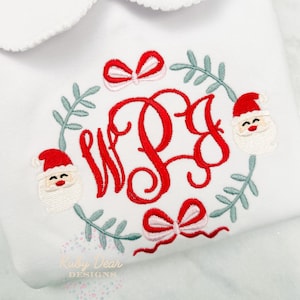 Puede incluir: Mono blanco con un diseño de corona de Navidad bordada en rojo y verde. La corona presenta un monograma "WPB", dos lazos rojos y dos caras de Papá Noel.