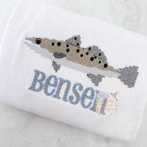 May include: White fabric with a cross-stitched fish design in shades of brown, gray, and black. The name "Benser" is embroidered in blue thread. A small logo with text and a hummingbird is in the bottom right corner.