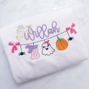 Puede incluir: Tela blanca con diseño bordado de Halloween. Presenta una pancarta con el nombre "Willah" en escritura morada, junto con un sombrero de bruja, un fantasma, una calabaza, arañas y lazos rosas. El diseño es probablemente para una prenda infantil.