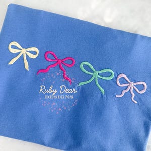 Puede incluir: Una tela azul con cuatro lazos bordados en colores pastel: amarillo, rosa, verde y lavanda. El texto "Ruby Dear Designs" está bordado debajo de los lazos en una fuente cursiva.