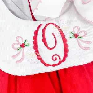 Puede incluir: Cuello blanco bordado con una letra O roja, lazos rosas y ramitas de acebo. El cuello está unido a un vestido de pana roja.