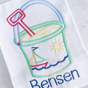 May include: White t-shirt with an embroidered beach bucket design. The bucket is filled with sand and a red shovel. A sailboat and sun are also embroidered. The name "Bensen" is embroidered in blue.