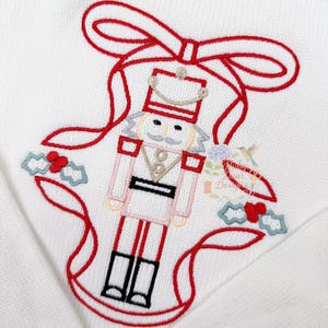 Könnte beinhalten: Weißer Strickstoff mit einem roten Stickdesign, das einen Nussknacker-Soldaten, eine rote Schleife und Stechpalmenblätter zeigt. Der Nussknacker trägt eine rosa Jacke und schwarze Stiefel. Das Design ist festlich und weihnachtlich.