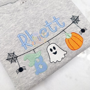 Puede incluir: Una camiseta gris claro de manga larga con el nombre "Rhett" bordado en azul claro. La camiseta presenta un diseño temático de Halloween con una calabaza, un fantasma, arañas y telarañas.