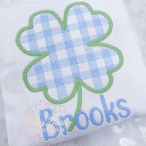 May include: A white t-shirt with a green four-leaf clover design with a blue and white gingham pattern inside. The name "Brooks" is embroidered in blue below the clover.