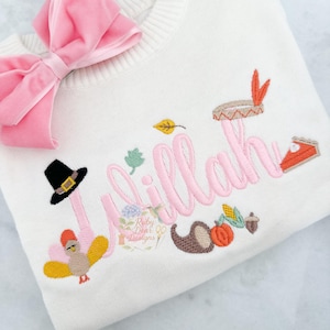 May include: Cream-colored sweater with pink embroidered name "Lillah" and Thanksgiving-themed designs. Features a pink bow, a turkey, a pilgrim hat, and a slice of pie. The sweater also has a leaf, corn, and acorn design.