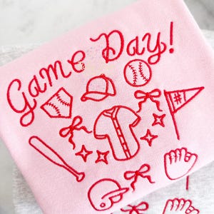 May include: Pink sweatshirt with red embroidery of baseball-themed illustrations and the text "Game Day!"
