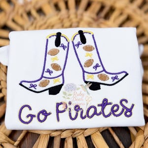 May include: White t-shirt featuring embroidered purple cowboy boots with footballs and bows. The text "Go Pirates" is embroidered in purple and gold. The shirt is displayed on a woven background.