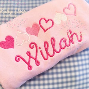 Könnte beinhalten: Rosaer Baby Strampler mit dem Namen "Willa" in rosaer kursivschrift gestickt und einer Reihe von fünf rosa und weißen Herzen.