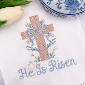 May include: White Easter-themed hand towel with an embroidered cross design. The cross is light brown, adorned with a blue bow and pink flowers. The words "He Is Risen" are embroidered in blue script below the cross.