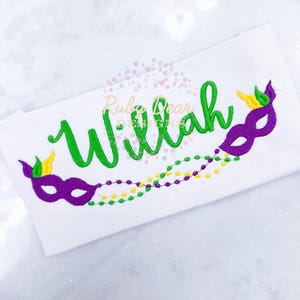 May include: A white fabric with the name "Willah" embroidered in green cursive lettering.  Two purple and gold Mardi Gras masks are embroidered on either side of the name, with green, gold, and purple beads embroidered below.