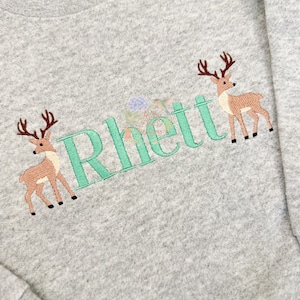Puede incluir: Una sudadera gris jaspeado con un diseño bordado de dos ciervos marrones con astas de color marrón oscuro que flanquean el nombre Rhett en letras grandes de color verde claro con serifa. Un pequeño detalle floral se ve sobre la letra h.