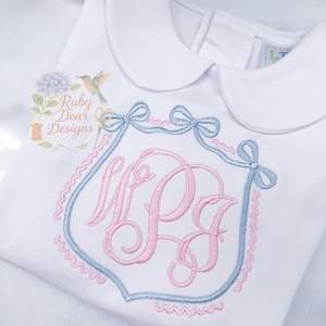 Puede incluir: Camisa blanca de manga larga con cuello Peter Pan. La camisa presenta un monograma bordado en azul claro y rosa dentro de un diseño de escudo, acentuado con lazos. El texto "Ruby Dear Designs" es visible.