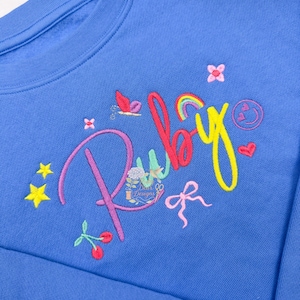 May include: A blue sweatshirt embroidered with the name "Ruby" in colorful script. The design includes a rainbow, stars, flowers, a bow, and cherries. The sweatshirt is a solid blue color.