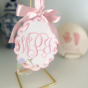 May include: A pink and white embroidered tag with a scalloped edge and a pink satin ribbon bow. The tag features the embroidered initials "WPS" in a script font. The tag is suspended from a gold-coloured metal stand.