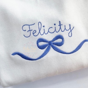 May include: White fabric with blue embroidery of the name "Felicity" and a blue ribbon bow.