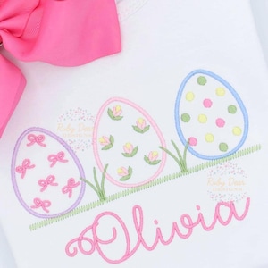 May include: A white onesie with a pink bow on top. The onesie has an embroidered design of three pastel Easter eggs with different patterns and the name "Olivia" underneath.
