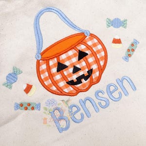 May include: A cream-colored tote bag featuring an embroidered orange and white gingham pumpkin basket with a blue handle. Black triangles form the eyes and mouth. Candy corn and wrapped candies are scattered around the pumpkin. The name "Bensen" is embroidered in blue.