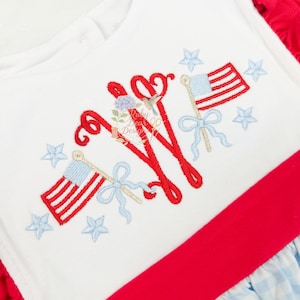 Pode incluir: Um vestido branco com um monograma vermelho bordado, duas pequenas bandeiras americanas com laços azul-claros e várias estrelas bordadas em azul-claro. A peça inclui uma cintura vermelha sólida e uma saia com padrão xadrez azul e branco.