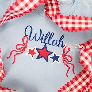 Może przedstawiać: Jasny błękitny śpioszek dla niemowlęcia z imieniem "Willah" wyszytym niebieskimi literami ozdobnymi. Trzy wyszyte gwiazdki w kolorze czerwonym, białym i niebieskim są ułożone w rzędzie pod imieniem. Wokół śpioszka owinięta jest czerwono-biała wstążka w kratkę.