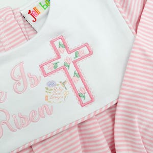 Può includere: Abito a righe rosa e bianche con colletto bianco. Il colletto presenta una croce ricamata rosa con fiori e le parole "He is Risen". L'abito ha maniche lunghe e una gonna a pieghe.