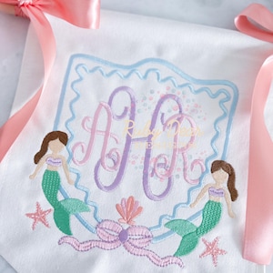 Könnte beinhalten: Weißes Stoffbanner mit Meerjungfrauen-Motiv. Mit einem Monogramm in Rosa und Lila, umrahmt von einem blauen, gewellten Rand. Zwei Meerjungfrauen, Seesterne und eine Schleife sind darunter gestickt. Rosa Bänder sind befestigt.